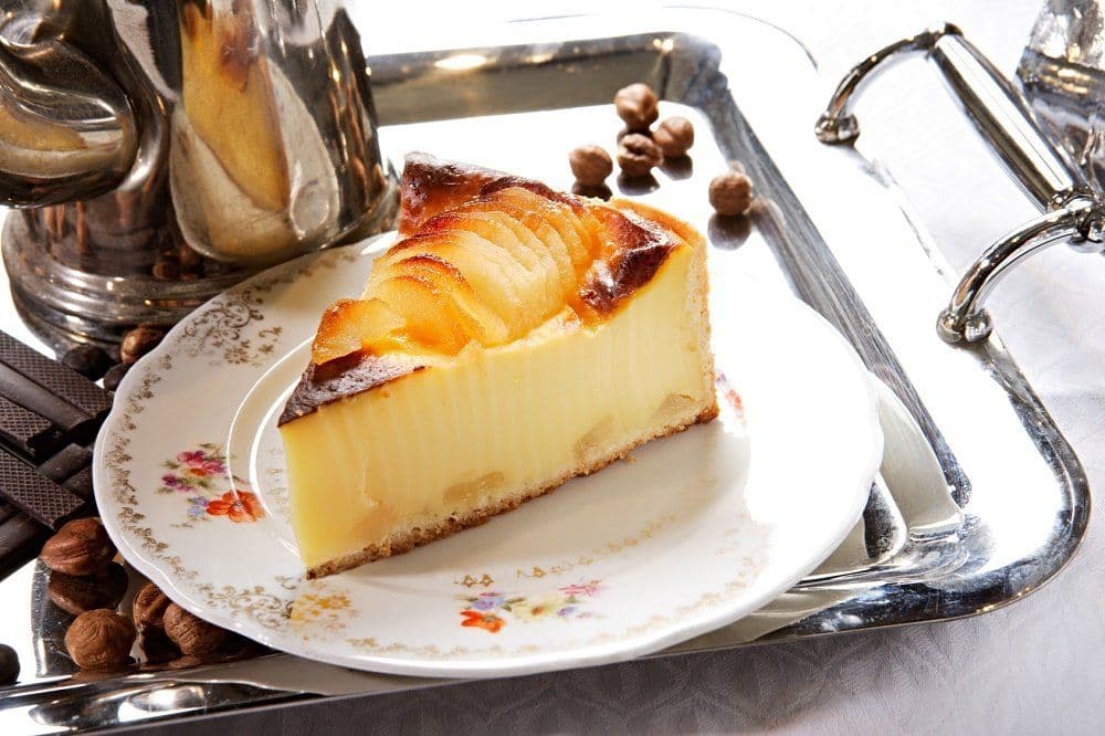 A delicious slice of cheesecake on a floral plate, surrounded by hazelnuts and a silver tray.